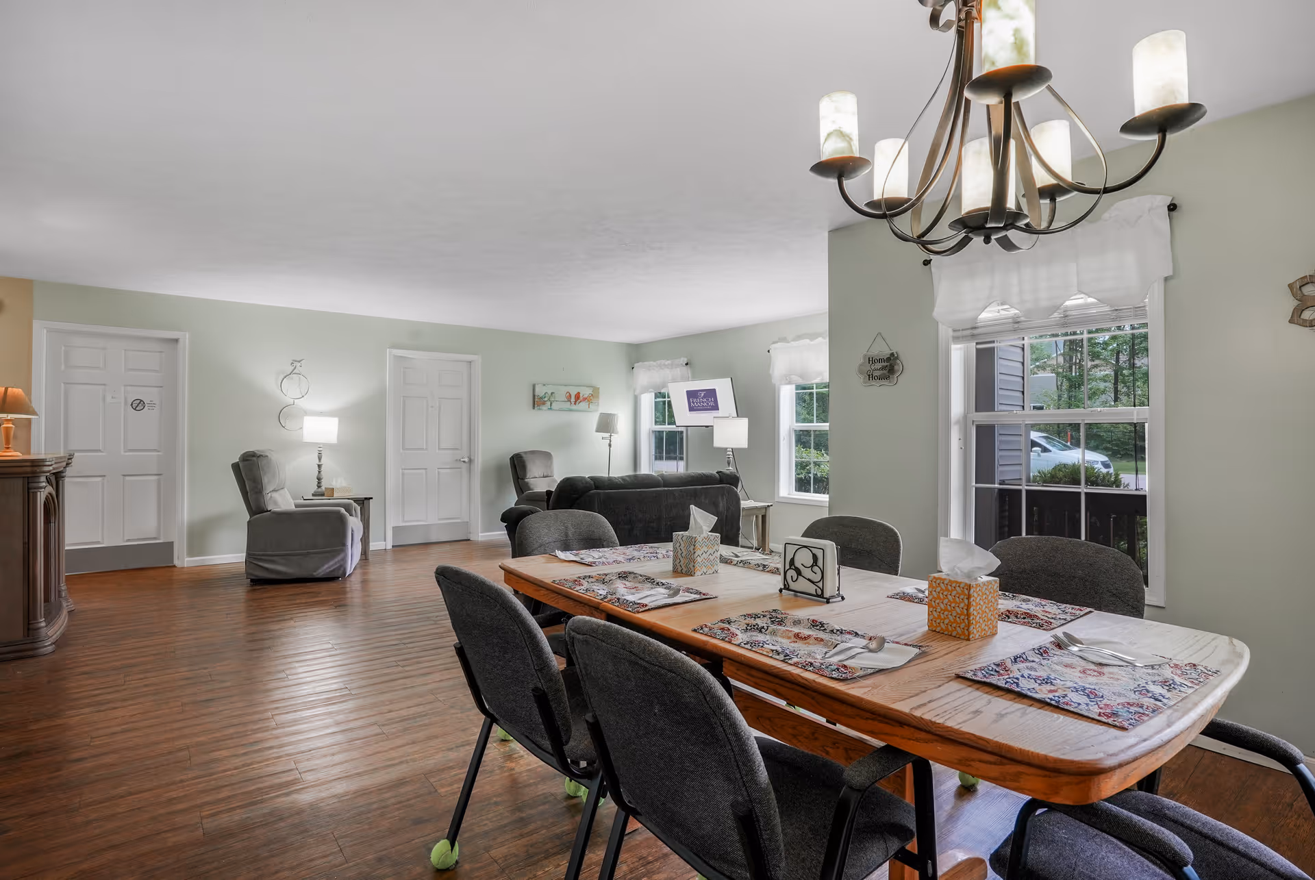 A spacious living and dining area with a wooden dining table set for six with placemats, tissue boxes, and utensils. The room has hardwood floors, light green walls, and multiple windows letting in natural light. There are two gray armchairs and a gray sofa in the living area, along with lamps and wall decorations. A chandelier with candle-style lights hangs above the dining table.