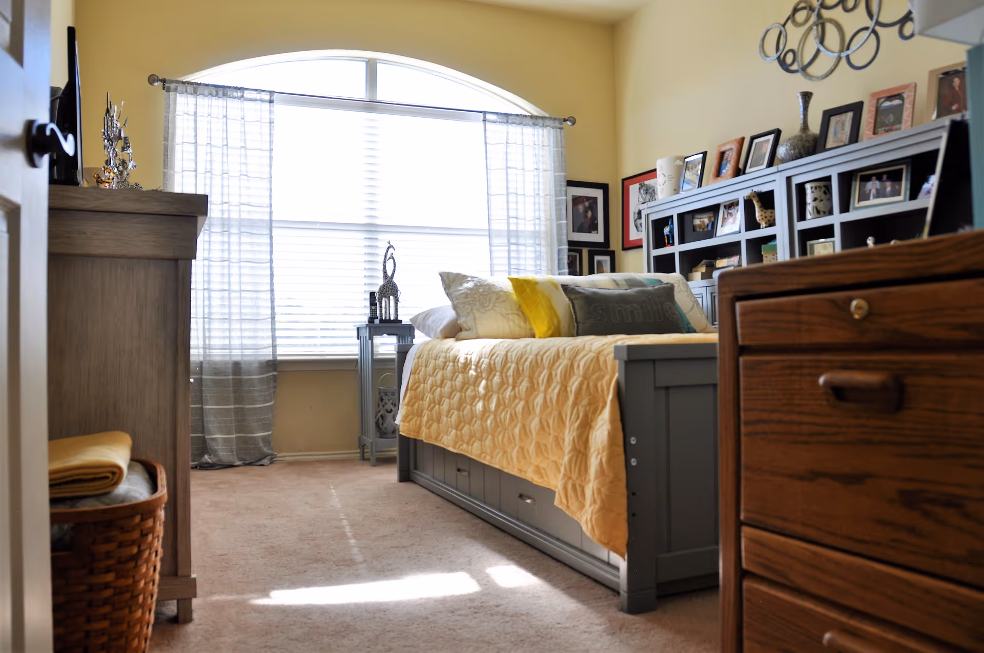 Sunny furnished bedroom with a yellow quilted bed, large arched window and shelving displaying framed photos.