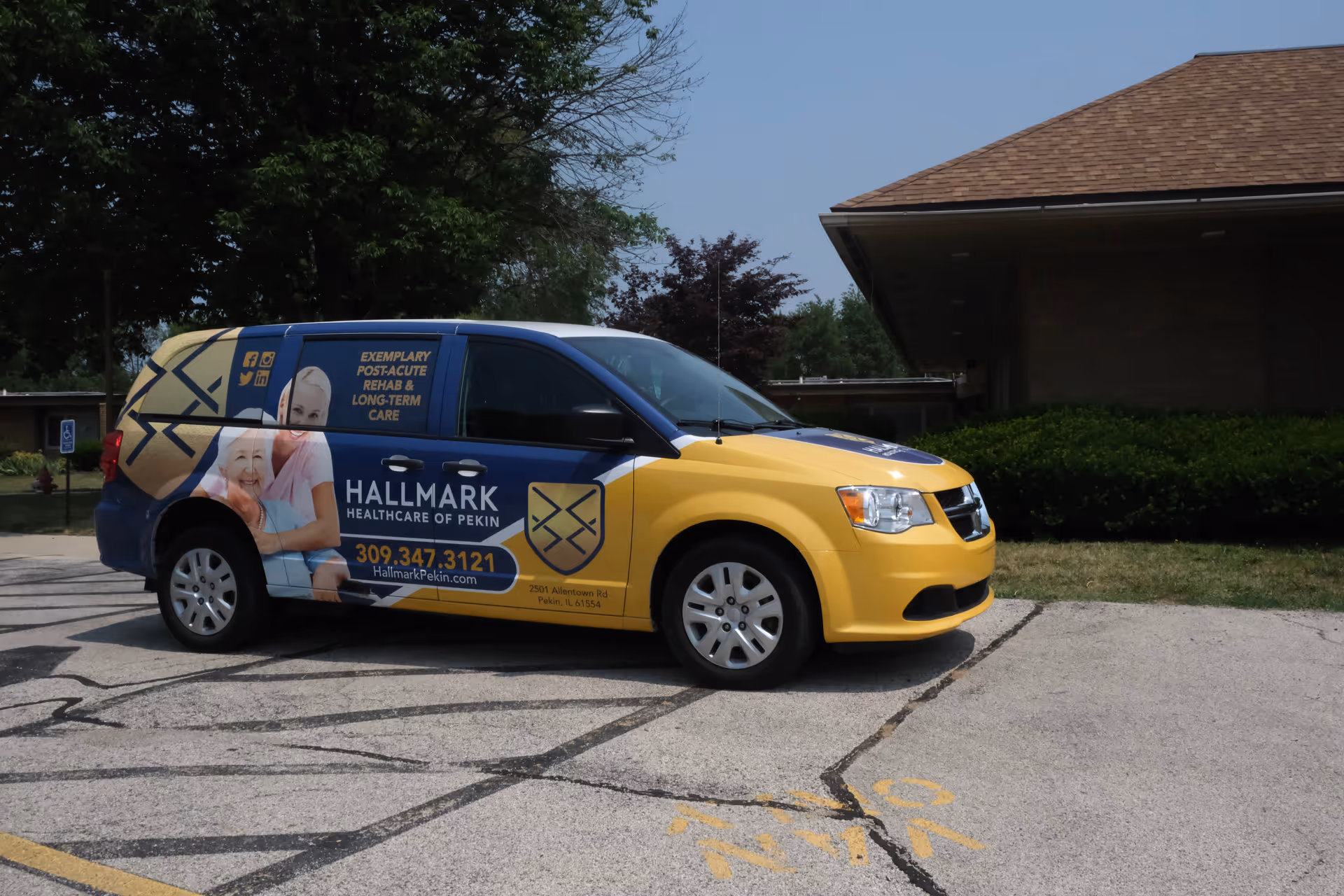 A yellow and blue van parked in a parking lot near a building. The van has signage for Hallmark Healthcare of Pekin, including images of a smiling elderly woman and a caregiver, contact phone number, and website. The background includes trees and part of a building with a brown roof.