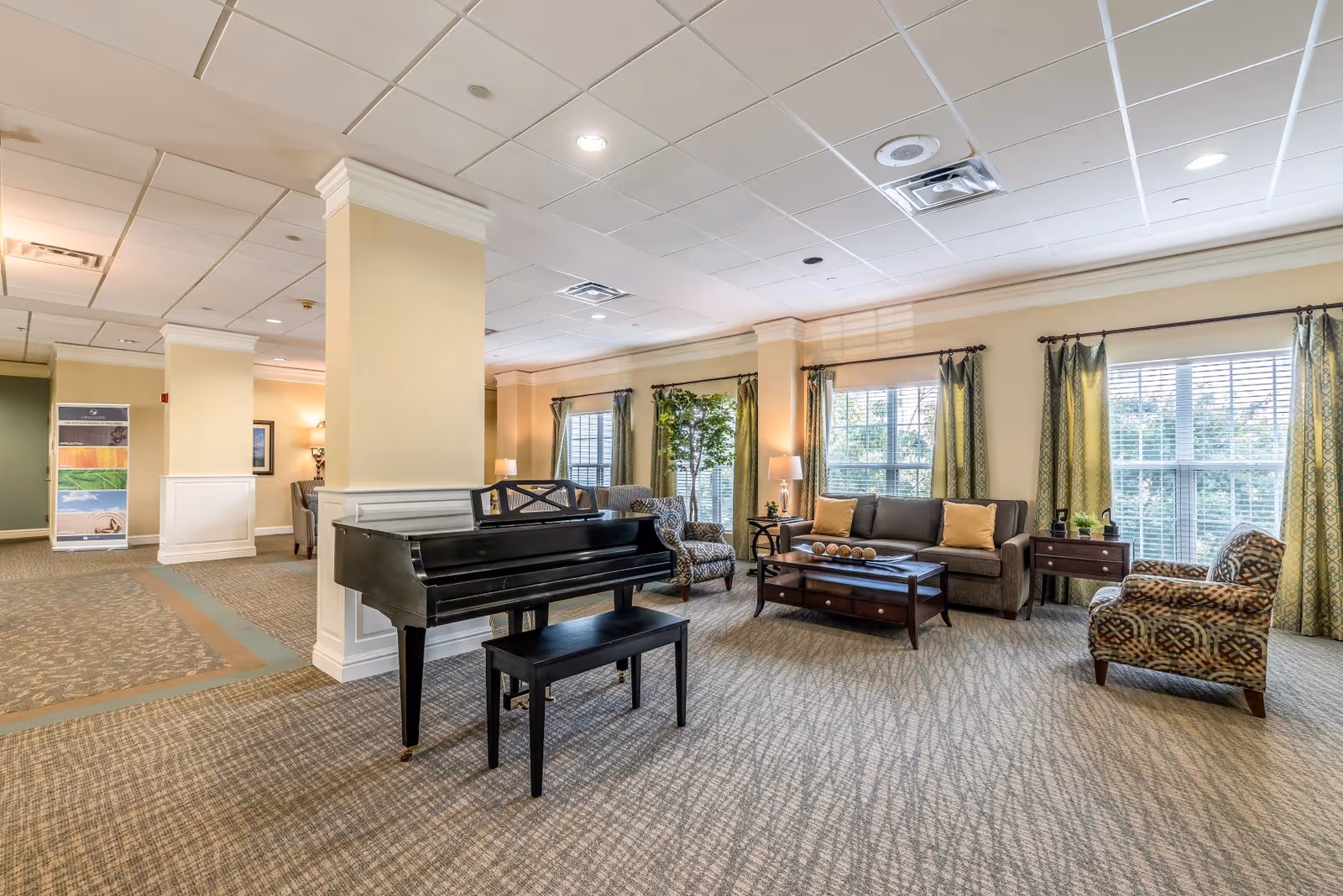 A spacious and well-lit living room area in a senior living facility featuring a black grand piano with a matching bench, a gray sofa with yellow cushions, two patterned armchairs, a wooden coffee table, side tables with lamps, large windows with green patterned curtains, and a potted plant near the window.