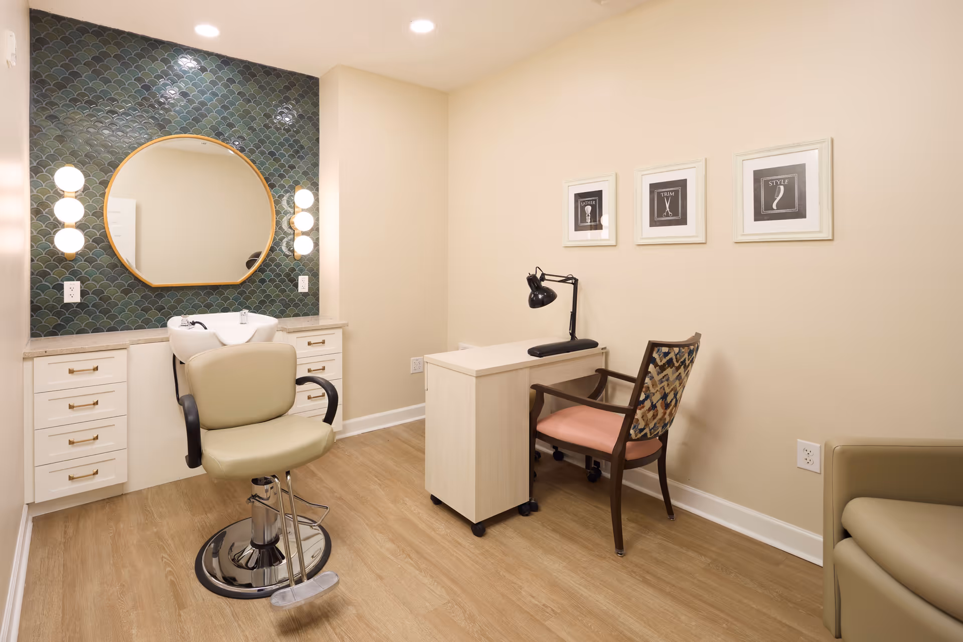 A small salon room with a beige salon chair in front of a white sink and a large round mirror with a green tiled wall behind it. To the right, there is a small desk with a black desk lamp and a wooden chair with a patterned backrest and pink seat cushion. The room has light wood flooring and beige walls with three framed pictures hanging above the desk.