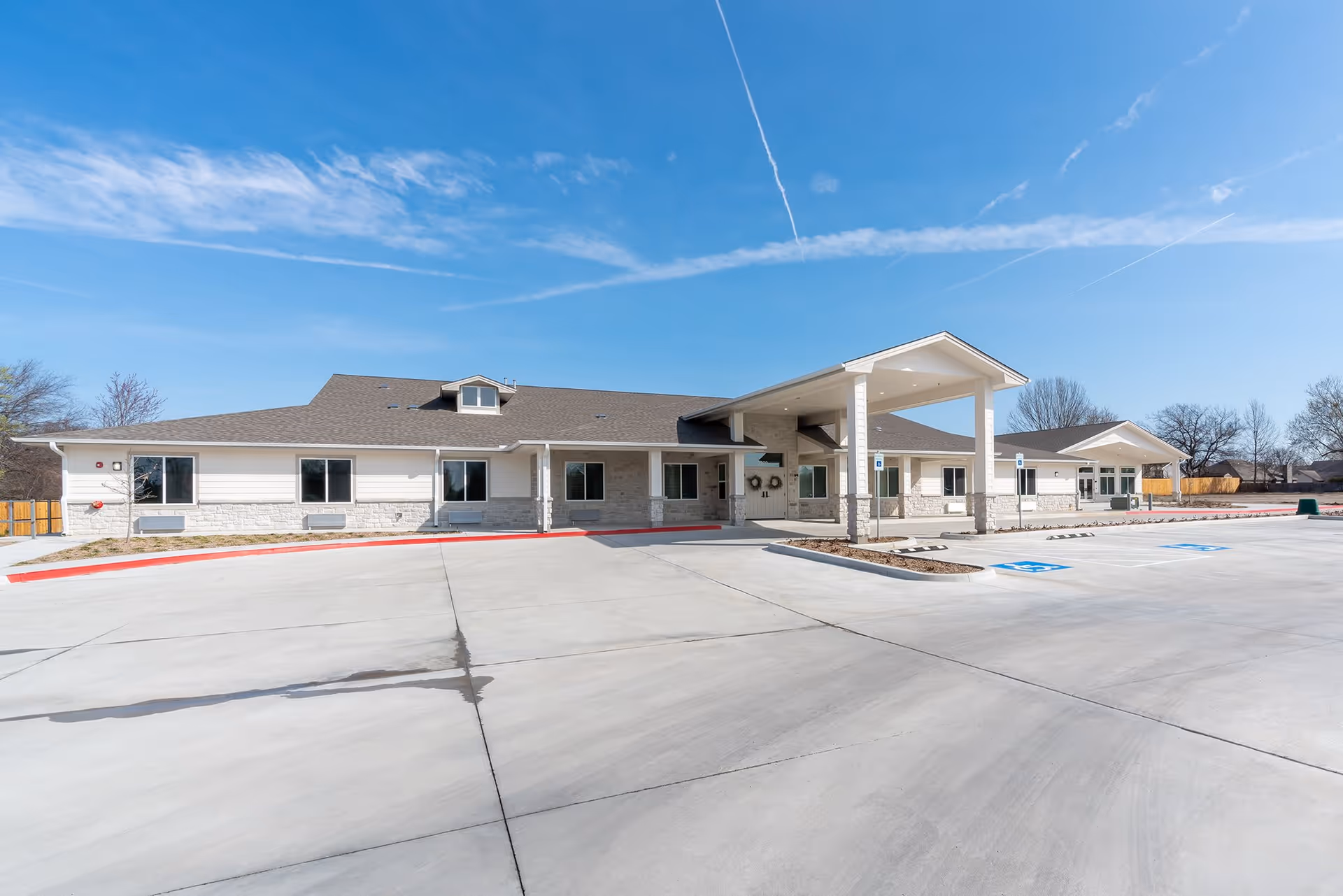 Exterior view of a single-story building with a covered entrance and multiple windows, surrounded by a large concrete parking lot with marked handicap parking spaces under a clear blue sky.