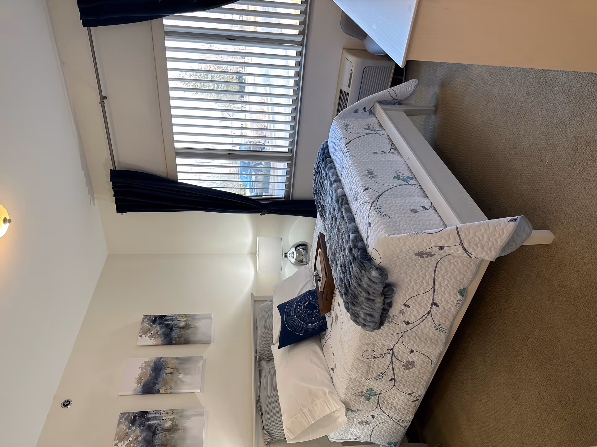 Sunlit, neatly made bedroom with a floral quilted bed, bedside lamp and tray, large window with blinds and dark curtains, and wall art.