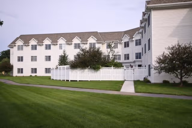Exterior view of a multi-story residential building with white siding and multiple windows, surrounded by green grass, trees, and a white fence enclosing a small garden area.