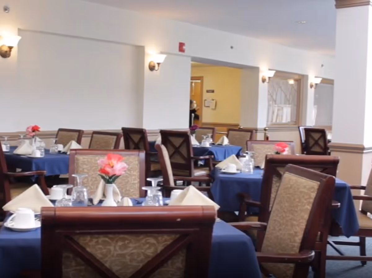 Dining room with multiple tables covered in blue tablecloths, each set with white napkins, cups, glasses, and small vases holding pink flowers. The room has beige walls with wall-mounted lights and wooden chairs with patterned upholstery.