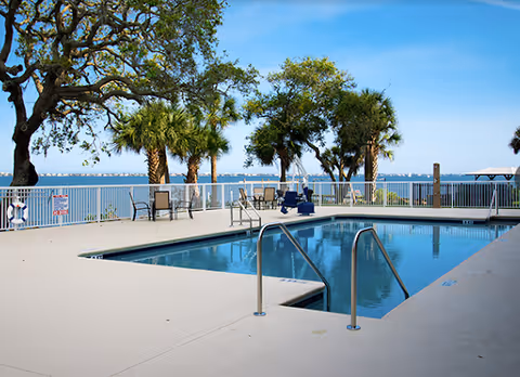 Outdoor swimming pool area with metal handrails, surrounded by a white fence. Several chairs and tables are placed near the fence, with palm trees and other greenery visible. In the background, there is a large body of water under a clear blue sky.