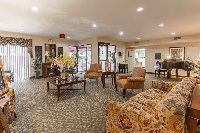 A spacious senior living facility common area with patterned carpet and beige walls. The room features a floral upholstered sofa, two striped armchairs, a glass coffee table with a flower arrangement, and a small side table with a lantern. In the background, there is a grand piano, framed artwork, and a fireplace with plants on either side. Large windows with vertical blinds and a glass door lead to an outdoor area. The ceiling has recessed lighting.