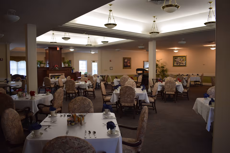 Spacious dining room with multiple tables set with white tablecloths and upholstered chairs under recessed ceiling lights.