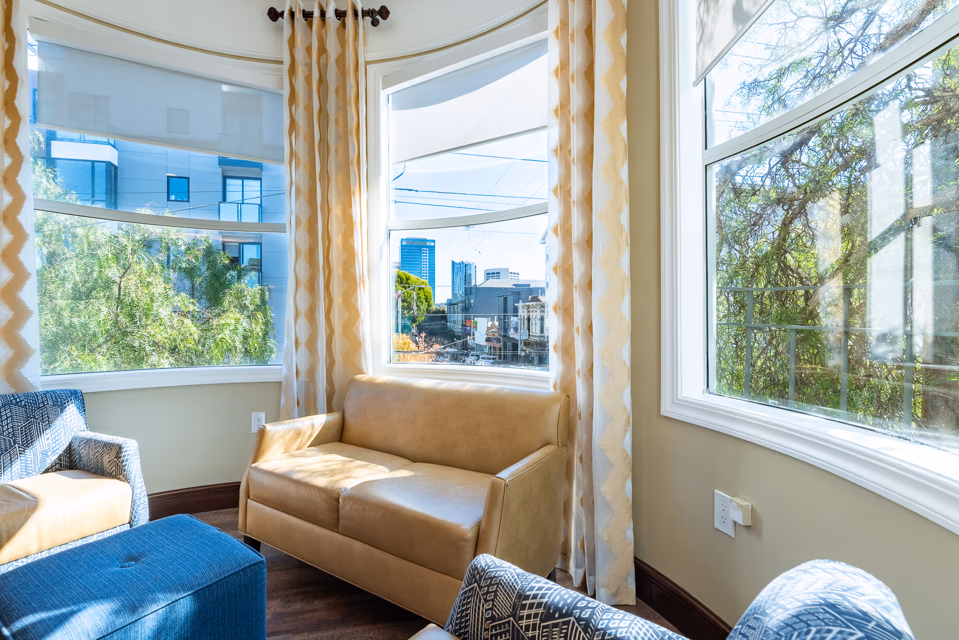 A bright sitting area with large curved windows showing an urban view outside. The room features a tan leather loveseat, two patterned armchairs, a blue ottoman, and beige curtains with a zigzag pattern. Sunlight fills the space, highlighting the wooden floor and light-colored walls.