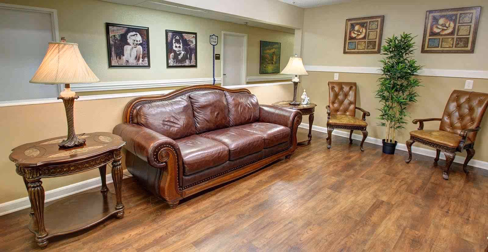 A cozy living room area with a brown leather sofa, two matching brown leather armchairs, two wooden side tables with lamps, a potted plant, and framed artwork on the walls. The room has wooden flooring and beige walls with white trim.