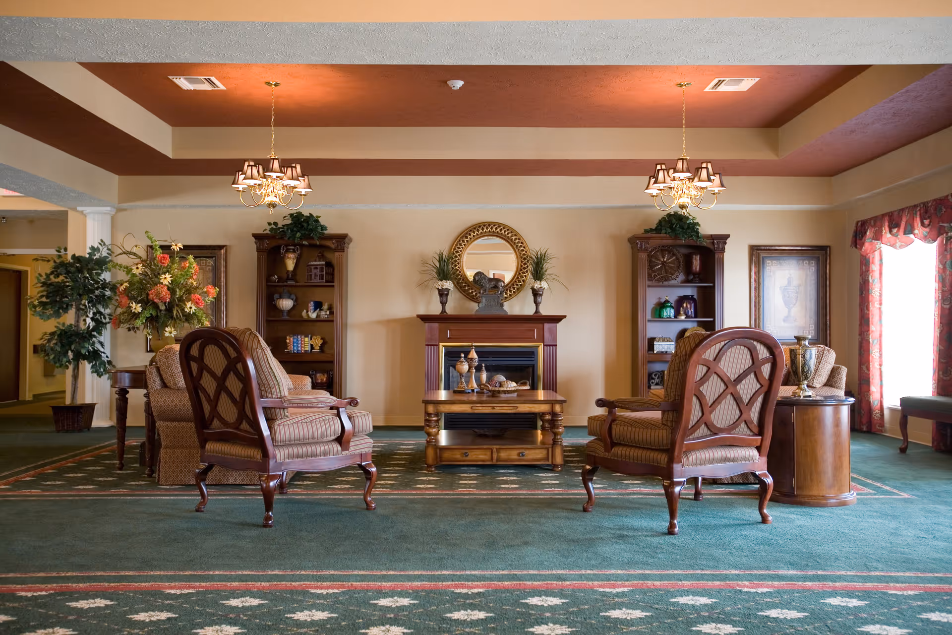 A cozy living room area with two wooden armchairs with striped cushions facing a wooden coffee table. Behind the table is a fireplace with a decorative mirror and plants on the mantel. Two wooden bookshelves flank the fireplace, each decorated with various ornaments. The room has green carpet with a patterned border, beige walls, and a ceiling with recessed lighting and two chandeliers. A large window with floral curtains allows natural light to enter the space.
