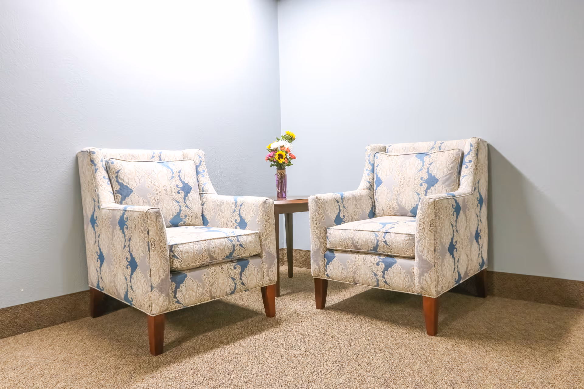 Two patterned armchairs with blue and beige designs are positioned facing forward with a small wooden table between them. On the table is a vase holding a colorful bouquet of flowers. The setting is in a corner with light gray walls and a brown carpeted floor.