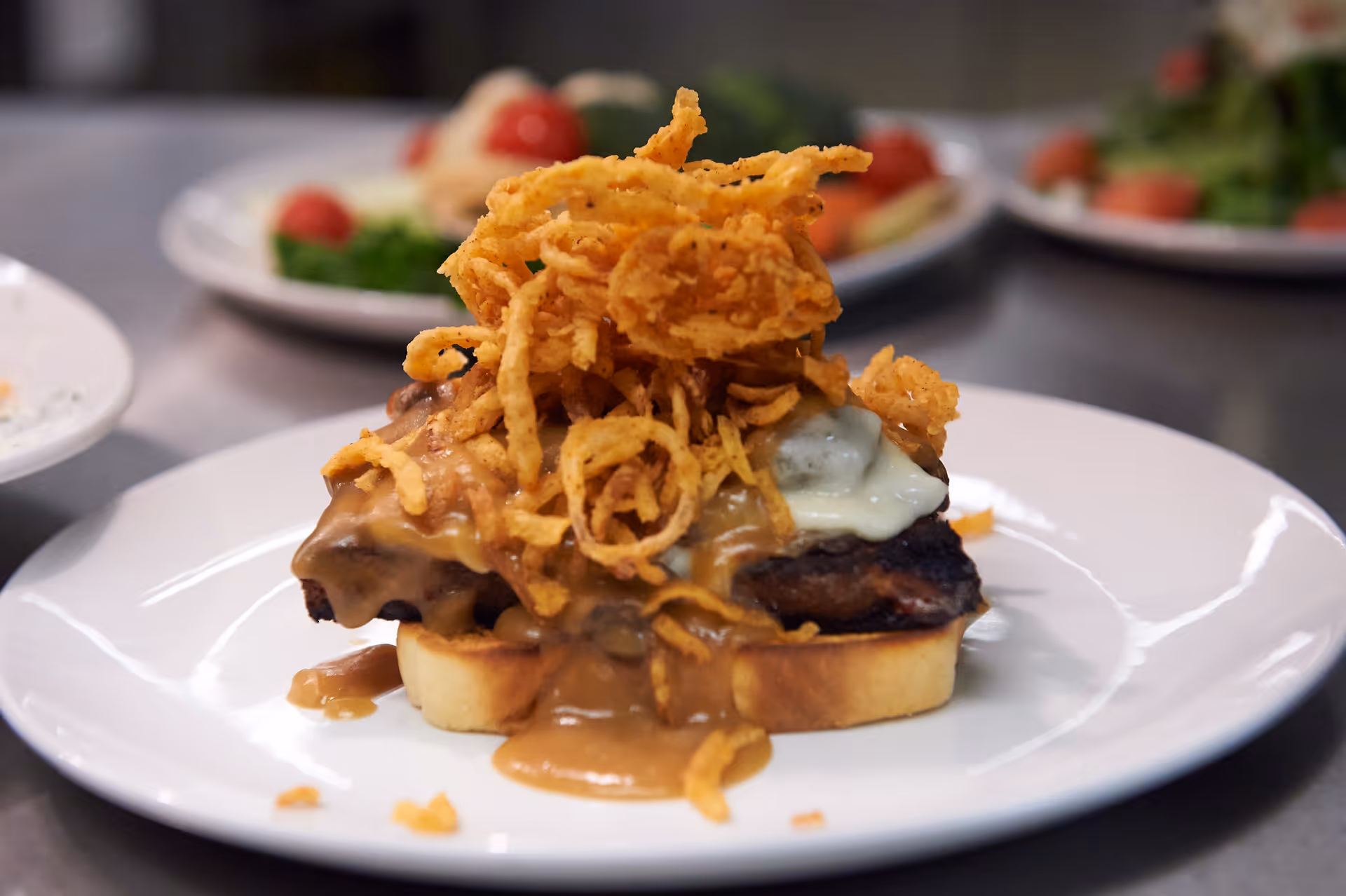 A close-up of a plated dish featuring a piece of meat topped with melted cheese, crispy fried onions, and brown gravy, served on a slice of toasted bread. In the background, there are blurred plates with salad and other food items.