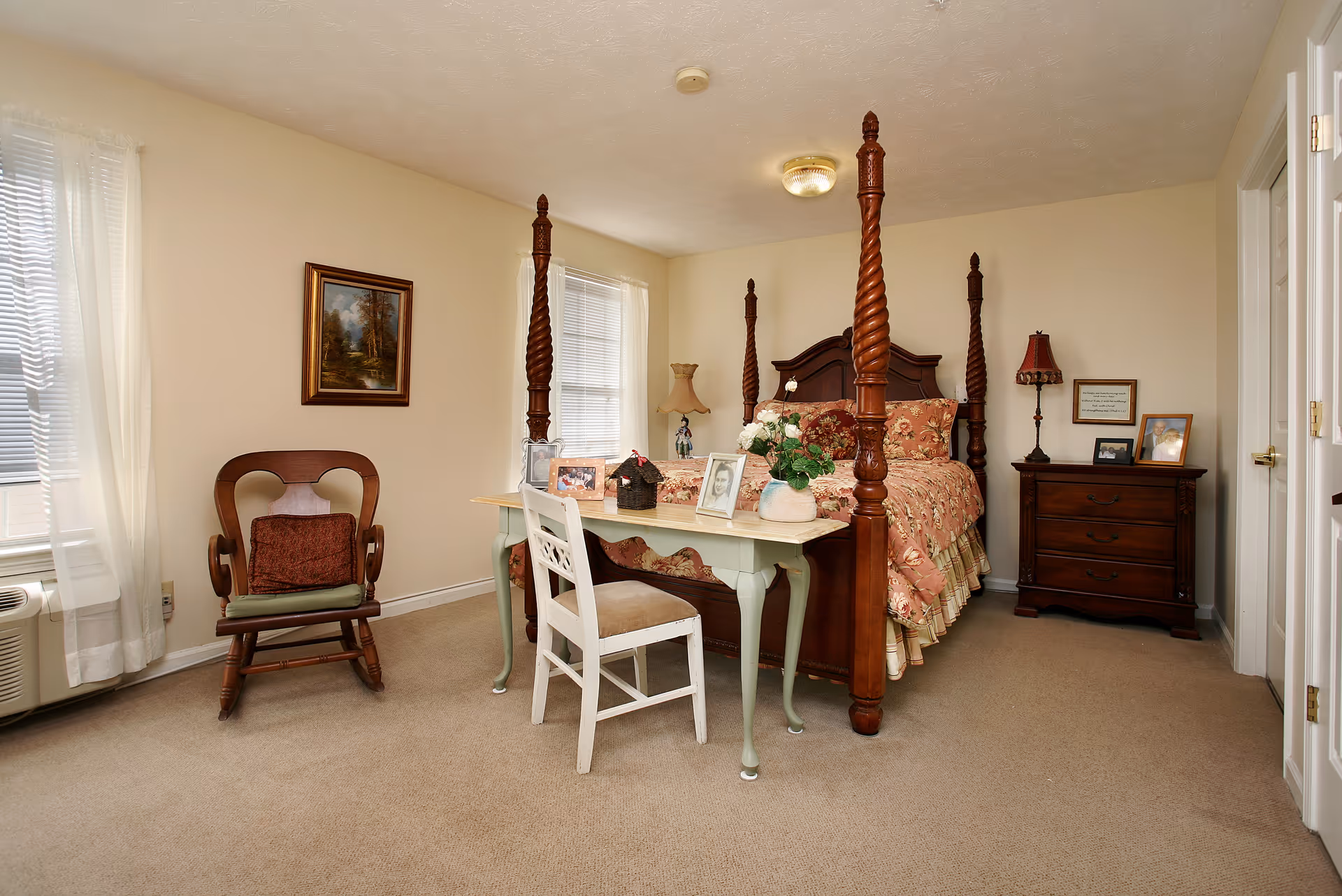 Well-lit traditional bedroom with a four-poster bed, bedside tables, a desk and chair, and a rocking chair by the window.