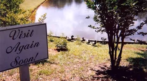 Sign reads Visit Again Soon beside grassy slope, trees, and a pond.