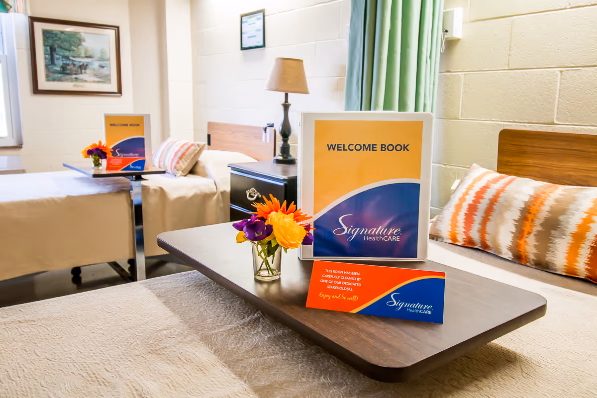 A bright and clean room with two beds, each with a bedside table and lamp. On the table in the foreground, there is a small vase with colorful flowers, a welcome book with the Signature HealthCARE logo, and a card indicating the room has been carefully cleaned. The room has light-colored walls, a framed picture, and a window with green curtains.