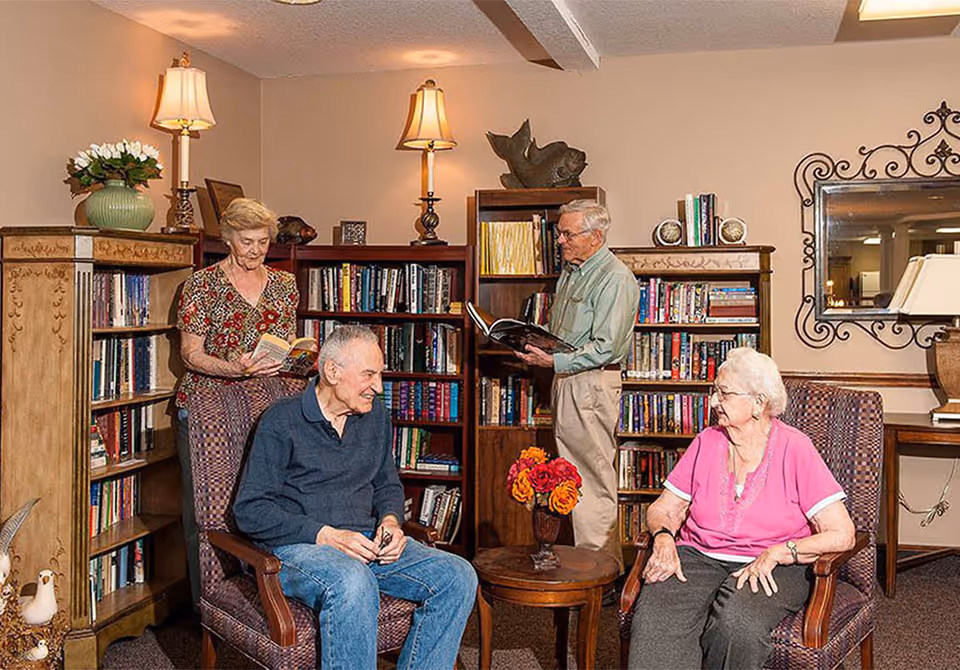 Four elderly residents sit and read or chat in a cozy common room with bookshelves, armchairs, and lamps.