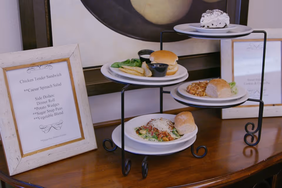 A three-tiered serving stand on a wooden table displaying plates of food including a sandwich with potato wedges and spinach, a plate with a dinner roll and beans, a salad with shredded cheese and a dinner roll, and a dessert plate with a slice of pie. Two framed menus are placed on the table beside the stand.