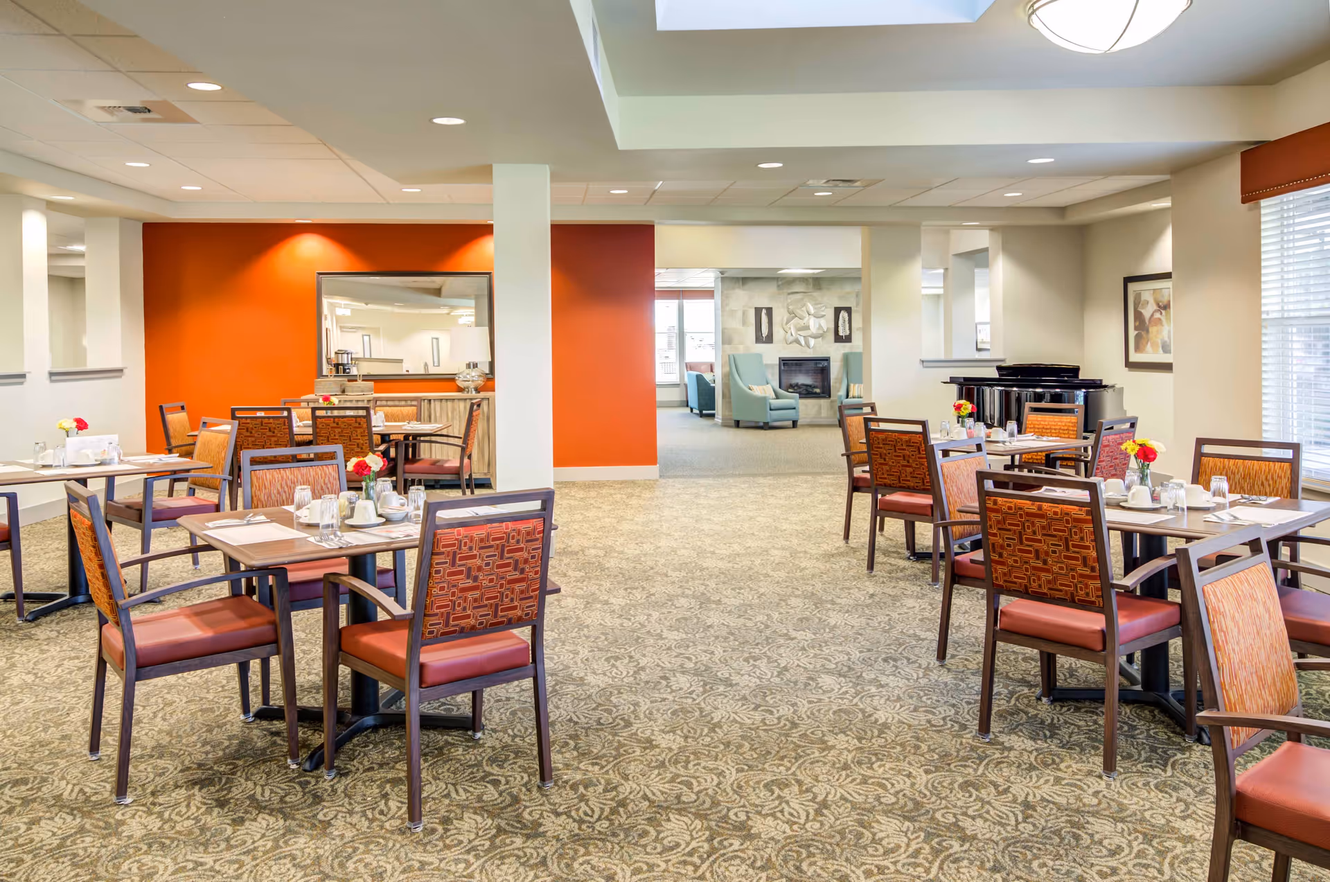 Bright dining room with multiple set tables and patterned chairs, leading to a seating area with a fireplace in the background.