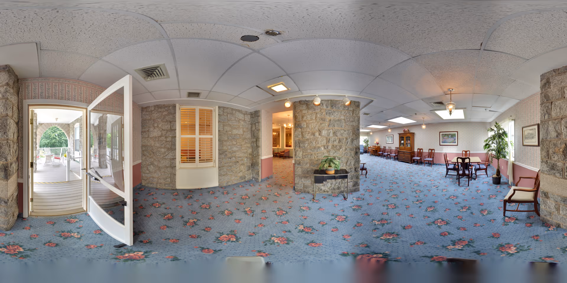 Interior view of a senior living facility lounge area with floral carpet, stone walls, and several wooden chairs and tables. There is a glass door leading to an outdoor patio with white railings and greenery visible outside. The room is well-lit with ceiling lights and natural light from windows.