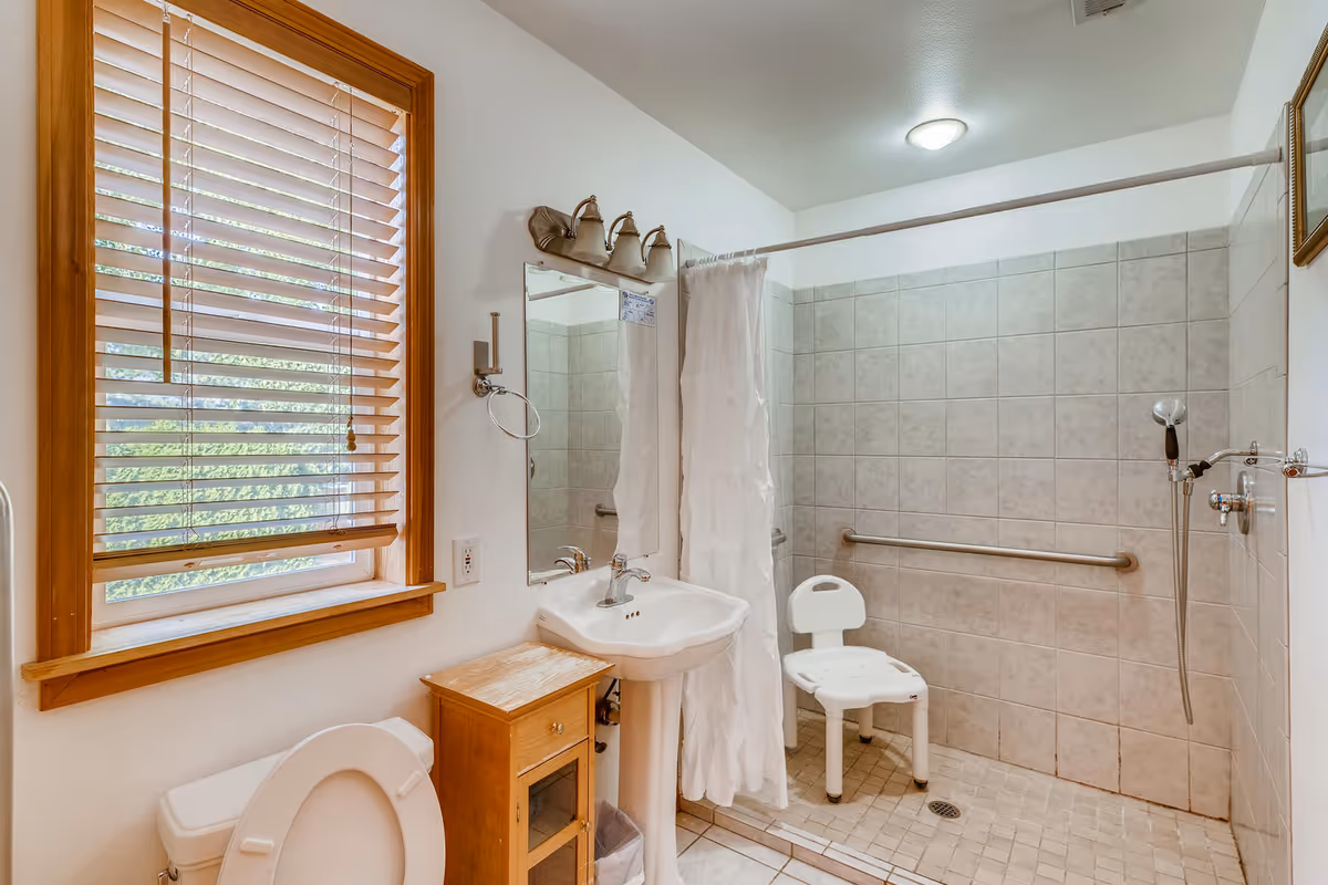 A bathroom with a window featuring wooden blinds, a white toilet with the lid open, a pedestal sink with a mirror and light fixture above it, a small wooden cabinet, and a walk-in tiled shower area with a white shower chair, grab bars, and a handheld showerhead.