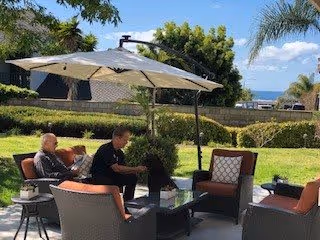 Outdoor seating area with four cushioned chairs and a glass-top table under a large patio umbrella. Two people are sitting and conversing in a garden setting with green grass, trees, and a view of the ocean in the background under a blue sky with some clouds.