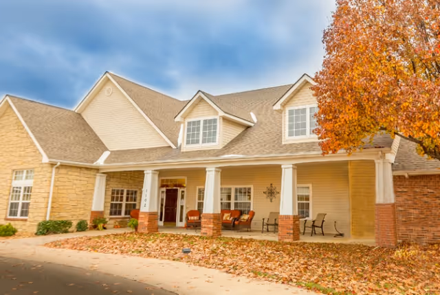 Front facade of a senior living building with a covered porch, chairs, and autumn leaves on the ground.