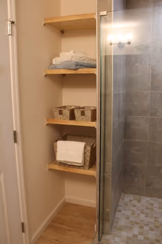 A bathroom corner featuring wooden shelves with folded towels and storage baskets next to a glass-enclosed tiled shower area.