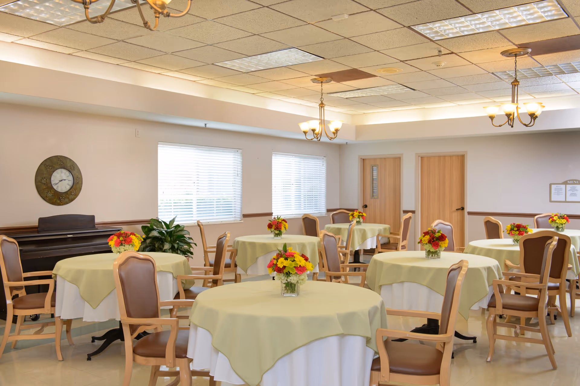 A dining room with multiple round tables covered with white and light green tablecloths, each table decorated with a small bouquet of red, yellow, and orange flowers. The room has wooden chairs with armrests, a piano against the wall, two windows with blinds, two wooden doors, and ceiling lights with chandeliers.