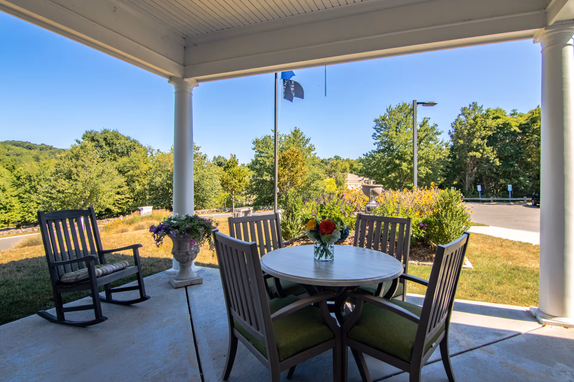 Covered outdoor patio area with a round table and four chairs, a rocking chair with a cushion, a flower vase on the table, and a potted plant near a white column. The background shows green trees, bushes, a flagpole with a flag, and a clear blue sky.