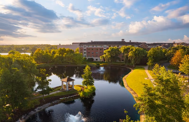 A scenic outdoor view of a senior living facility named Sedgebrook featuring a large pond with fountains, a small gazebo on a narrow strip of land, walking paths, lush green trees, and a multi-story brick building in the background under a partly cloudy sky.