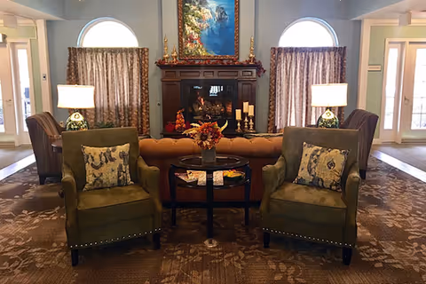 A cozy living room area with two green armchairs featuring patterned cushions, a round black coffee table with a floral arrangement, and a brown tufted sofa. Behind the sofa is a wooden fireplace mantel decorated with candles and a framed painting of a waterfall. Two windows with curtains flank the fireplace, and there are two table lamps on side tables next to the armchairs. The room has a warm, inviting atmosphere with carpeted flooring.