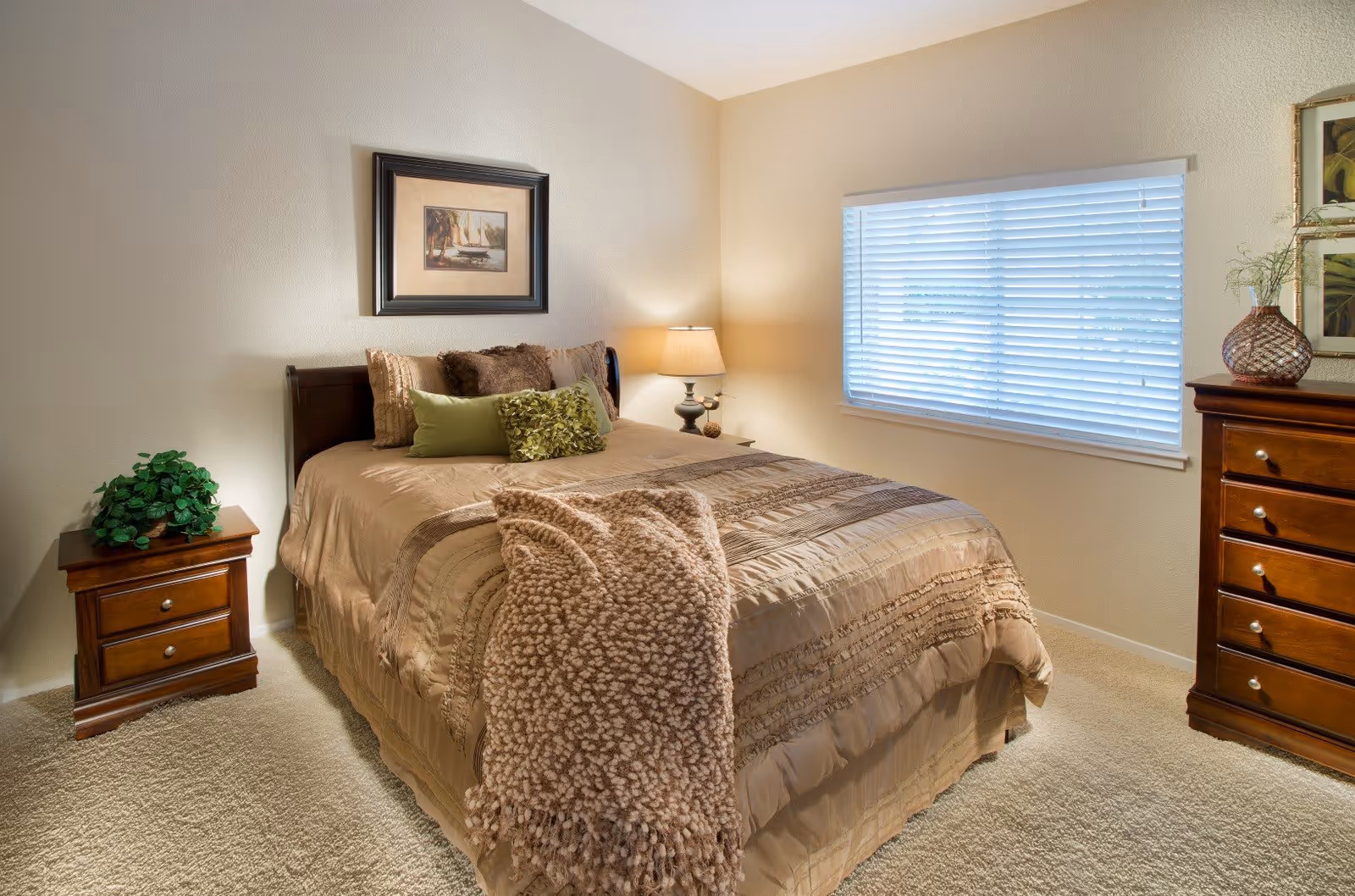 A cozy bedroom with a neatly made bed featuring beige bedding and multiple decorative pillows. A textured beige throw blanket is draped over the foot of the bed. There is a wooden nightstand with a green plant on the left side and a table lamp on the right side. A window with white blinds lets in natural light, and a wooden dresser with decorative items is on the right. A framed picture hangs above the bed on the light-colored wall.