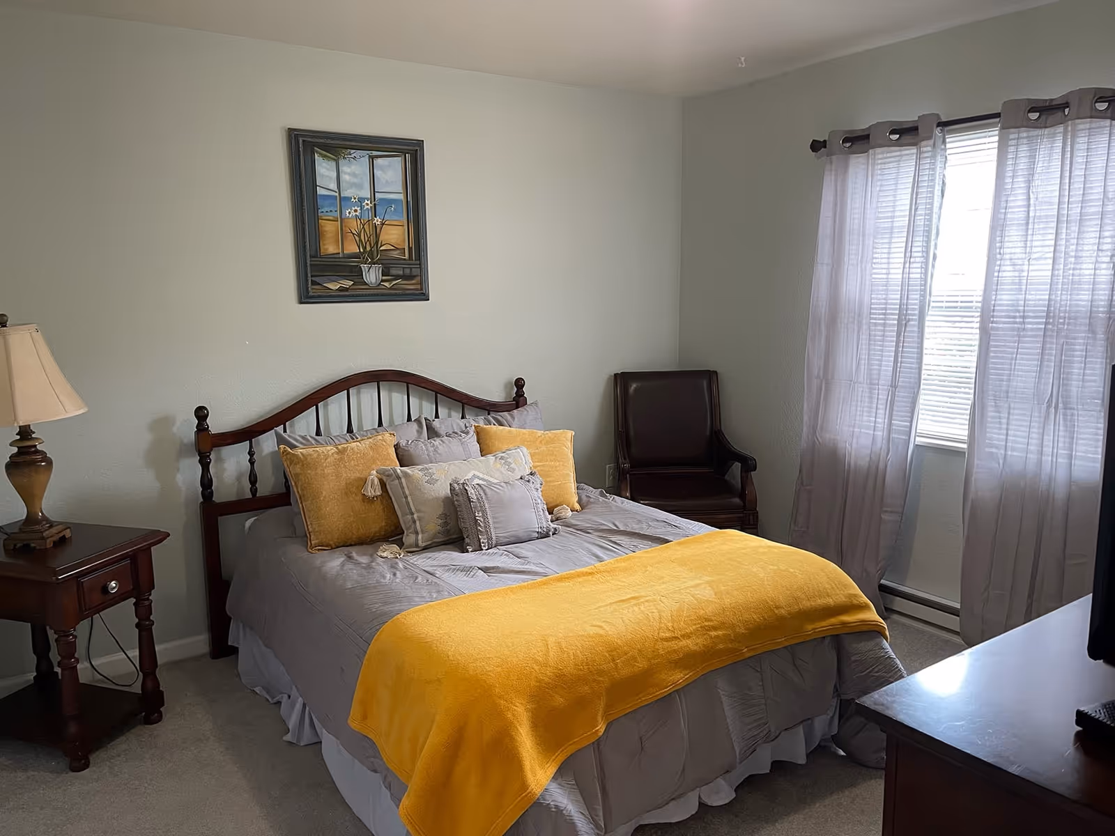 Sunlit bedroom with a wooden headboard bed dressed in gray linens and yellow accents, a nightstand with lamp, an armchair, and a window with sheer curtains.