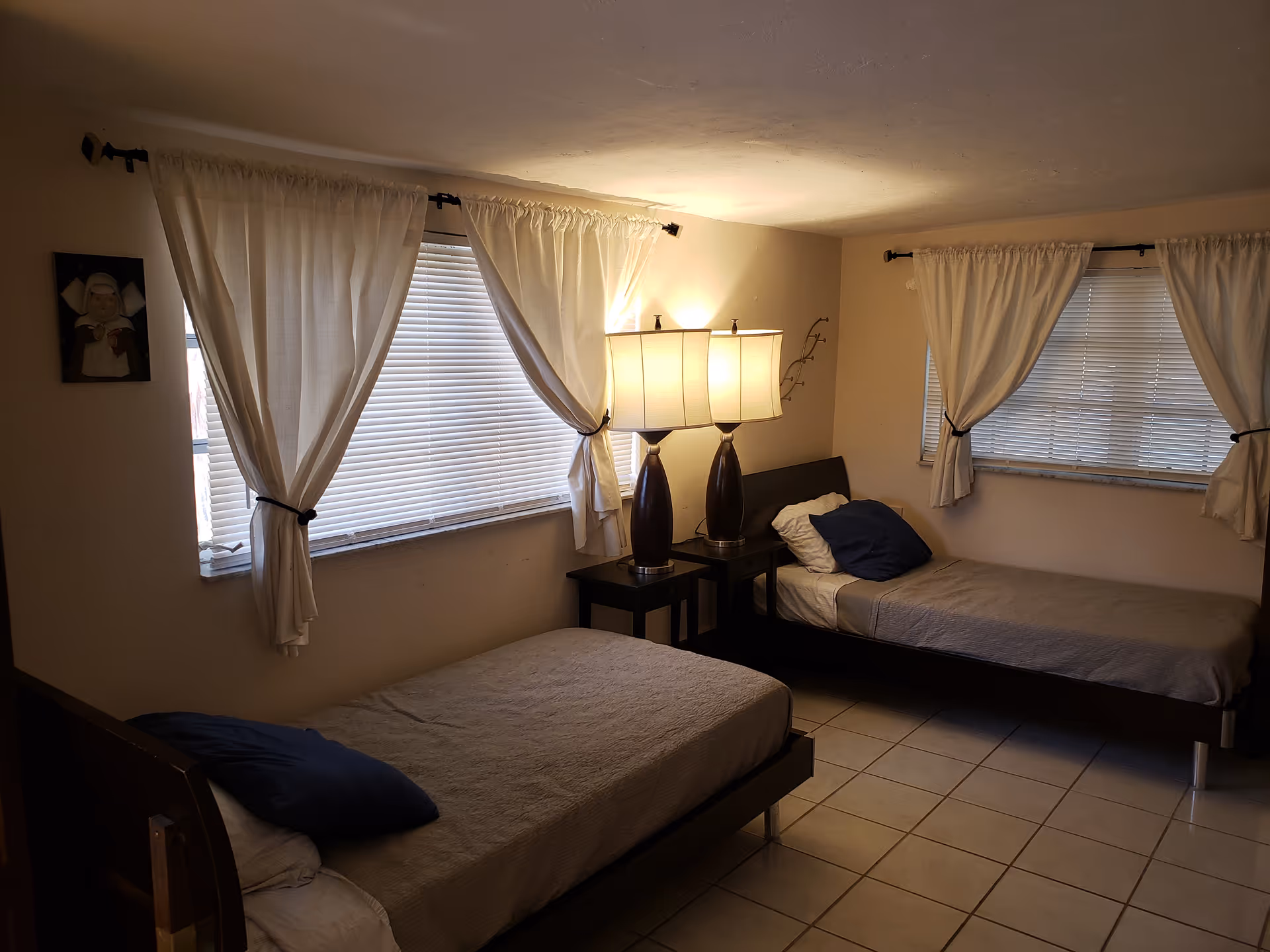 A simple bedroom with two single beds placed against adjacent walls. Each bed has a pillow and a beige bedspread. Between the beds is a small dark wooden nightstand with two matching table lamps that have beige lampshades. The room has tiled flooring and two windows with white blinds and white curtains tied back. A small framed picture hangs on the wall near one window.