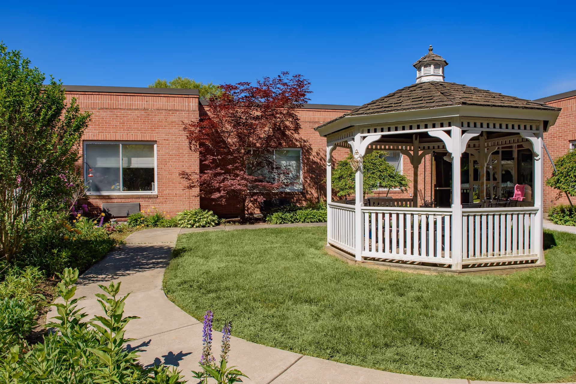 Outdoor courtyard area with a white wooden gazebo on a grassy lawn, surrounded by a curved concrete walkway, green shrubs, and trees. The background shows a brick building with windows under a clear blue sky.