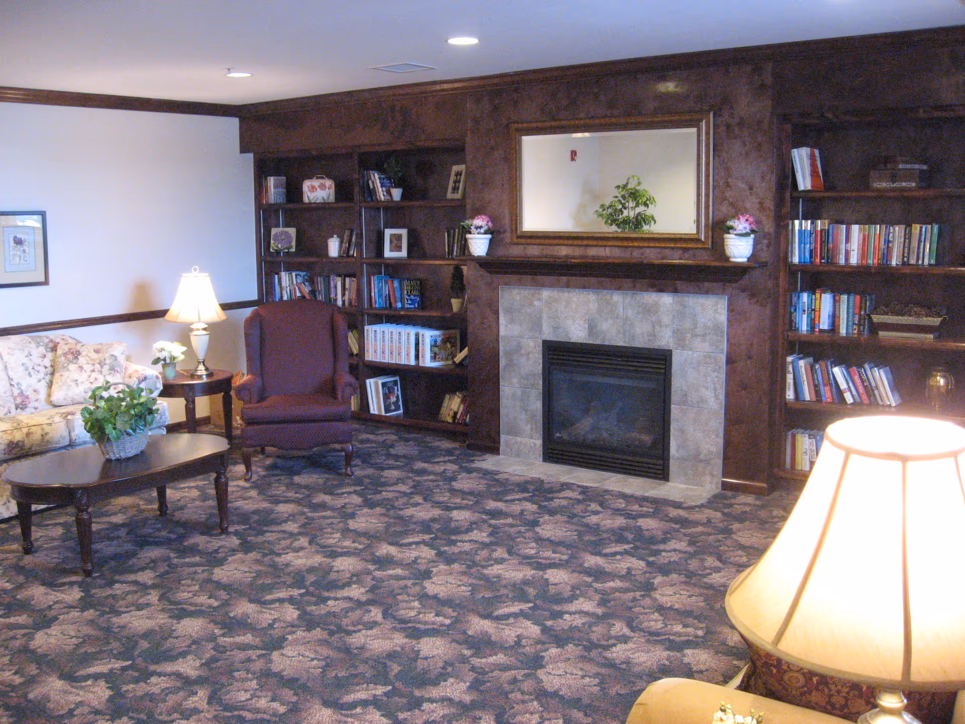 A cozy senior living facility common area with a floral patterned carpet, a floral sofa, a burgundy armchair, a wooden coffee table with a plant, two wooden bookshelves filled with books and decorative items, a fireplace with a tiled surround, and a large mirror above the fireplace. There are two table lamps providing warm lighting.