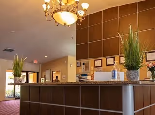 Interior view of a reception area with a wooden front desk adorned with two potted plants. Behind the desk are framed certificates on the wall. A chandelier hangs from the ceiling, and a doorway leading to another room is visible in the background.