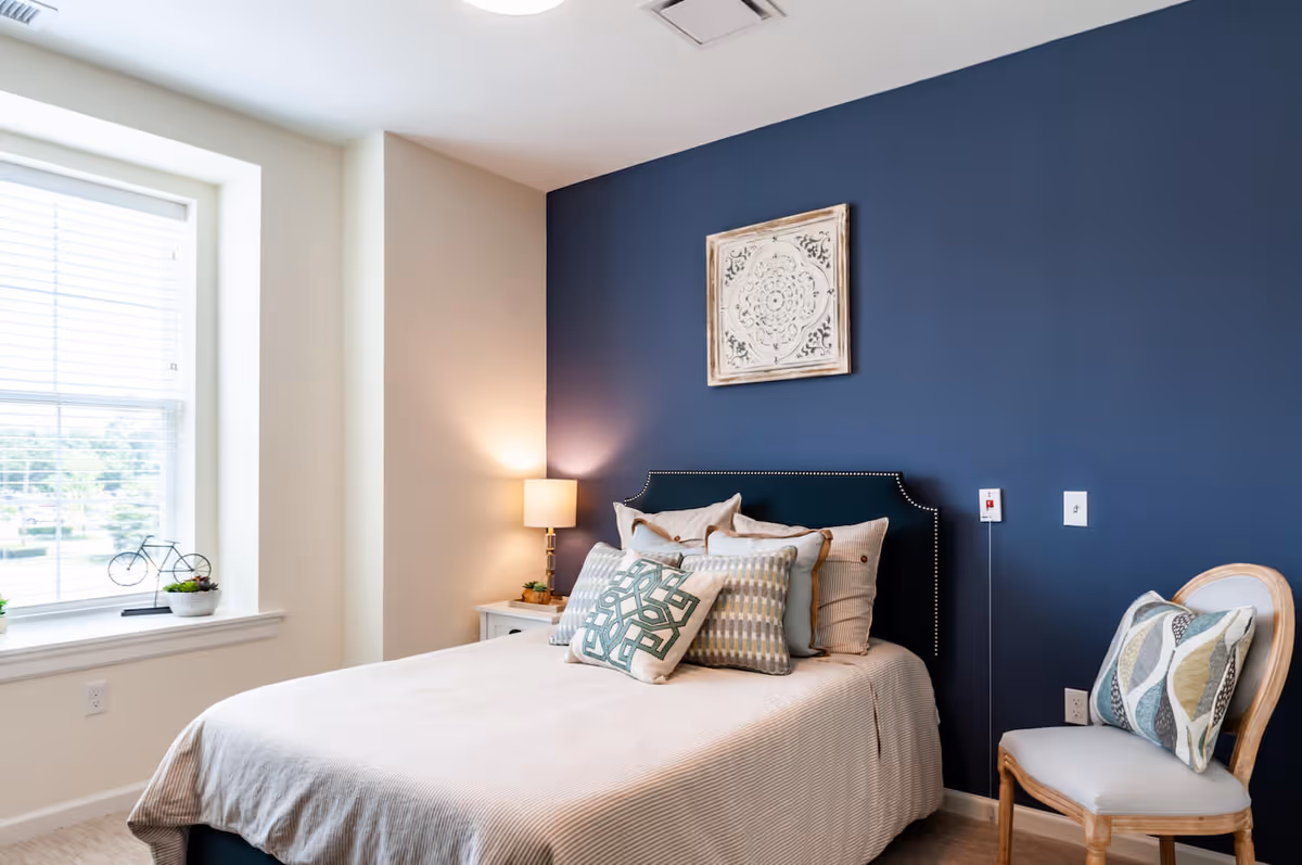 Bright bedroom with a bed against a navy accent wall, patterned pillows, a bedside table with lamp, and a window.