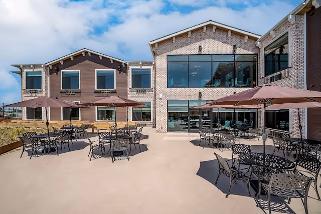 Outdoor patio area of a senior living facility with multiple black metal tables and chairs, each shaded by large brown umbrellas. The building in the background has large windows and a combination of light brick and brown siding under a partly cloudy blue sky.