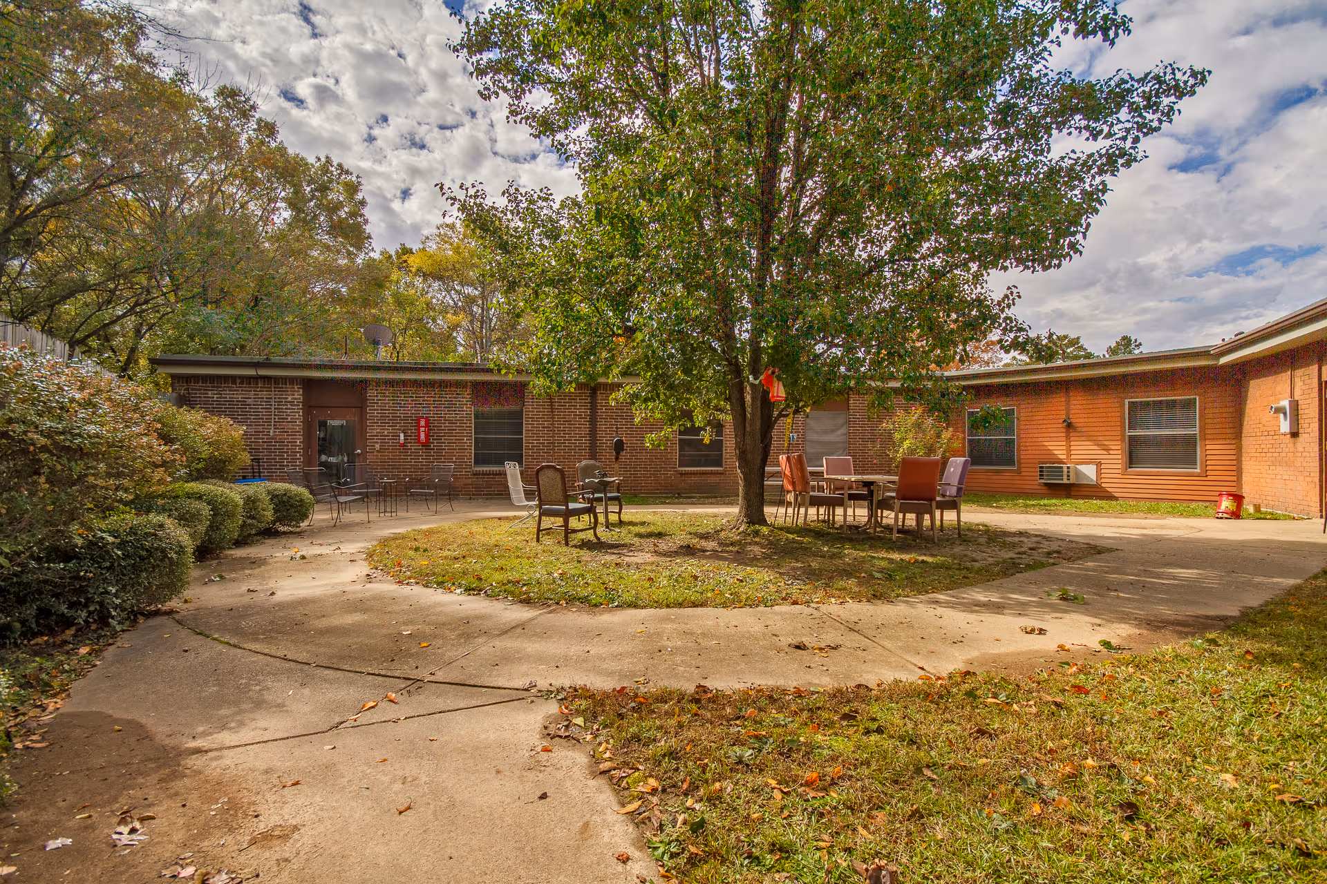 Outdoor courtyard area with a large tree in the center surrounded by a circular concrete walkway. Several chairs and tables are placed around the tree. The courtyard is enclosed by a single-story brick building with windows and doors. Bushes and trees with autumn foliage surround the area under a partly cloudy sky.