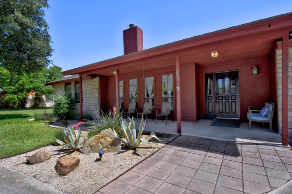Front exterior view of a single-story building with a covered porch featuring several chairs. The building has red siding and stone accents, with a paved walkway leading to the entrance. There are desert plants and rocks in a landscaped area near the walkway, and trees and grass in the background under a clear blue sky.