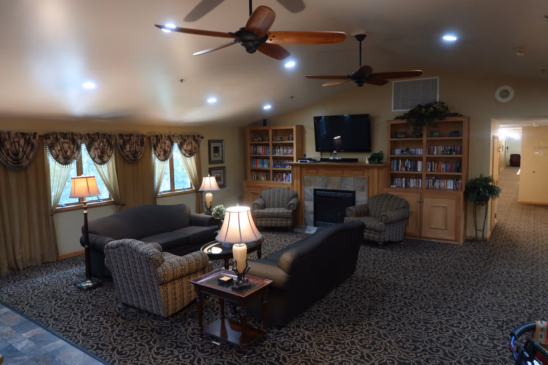 A cozy senior living facility common area with two dark sofas, two patterned armchairs, three table lamps, a fireplace with a mounted TV above it, and two wooden bookshelves filled with books. The room has patterned carpet, ceiling fans, and windows with draped curtains letting in natural light. A hallway is visible to the right.