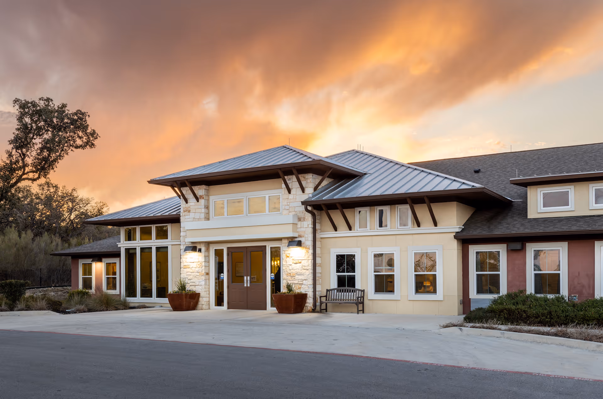 Exterior view of a senior living facility building at sunset with a metal roof, large windows, double entrance doors, potted plants, and a bench near the entrance.