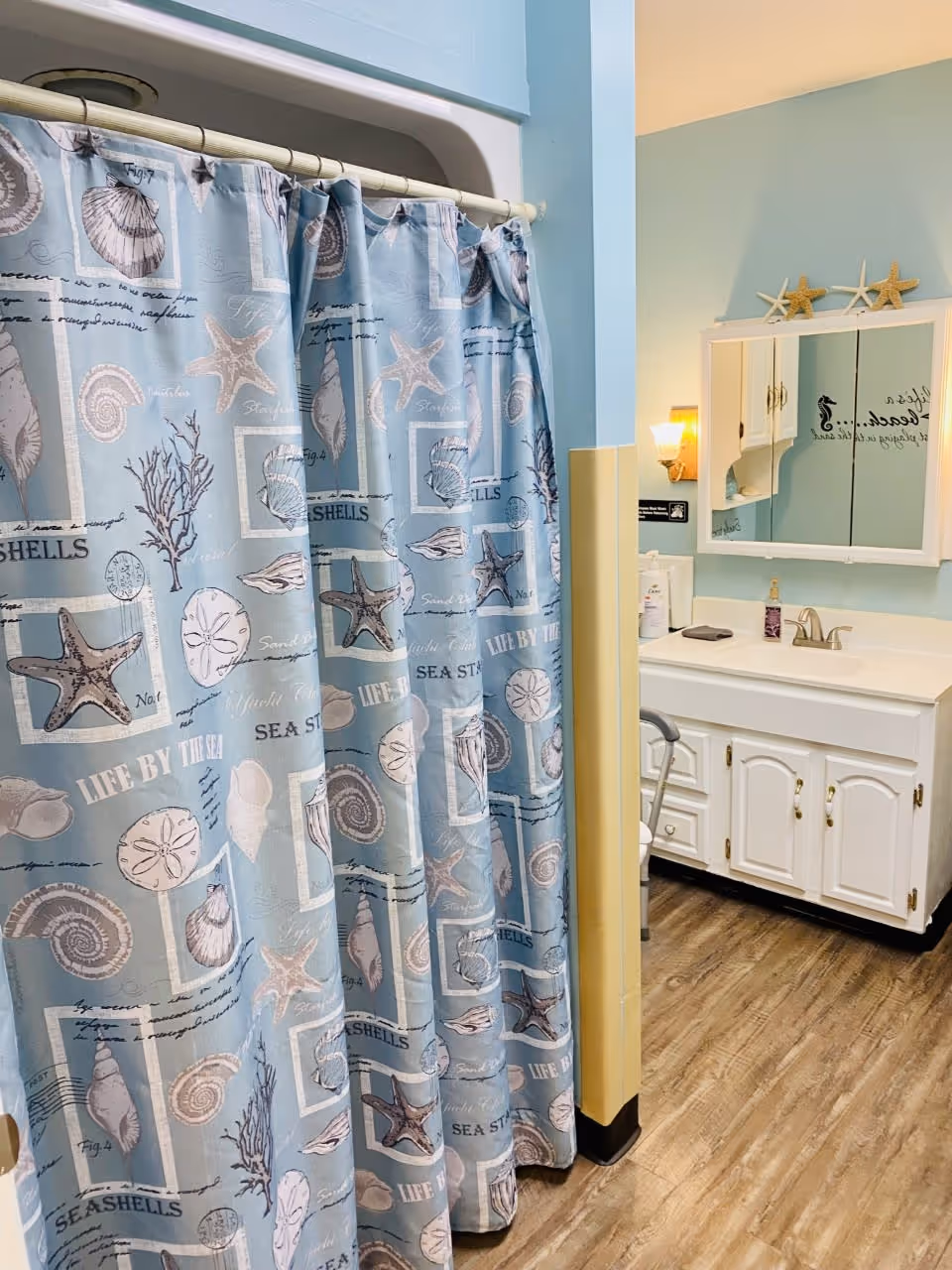A bathroom with a shower curtain featuring seashell and starfish patterns in blue tones. To the right, there is a white vanity with a sink, a mirror cabinet above it, and a wall-mounted light fixture. The walls are painted light blue and the floor has wood-like vinyl flooring.