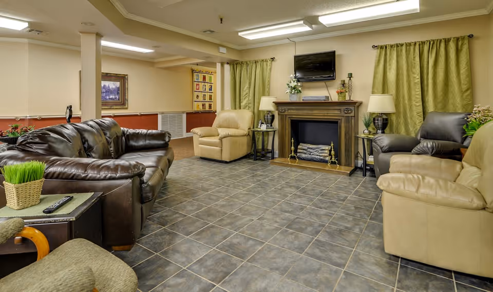 A cozy living room area in a senior living facility featuring a dark brown leather sofa, beige and dark brown armchairs, a wooden fireplace with decorative logs, a flat-screen TV mounted above the fireplace, green curtains, and a tiled floor. There are side tables with lamps and plants, and framed artwork on the walls.