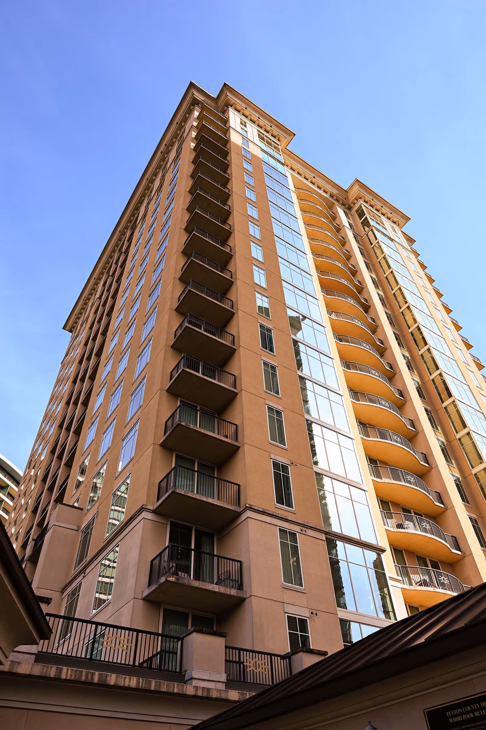 Low-angle view of a tall tan residential high-rise with stacked balconies and large reflective windows against a blue sky.