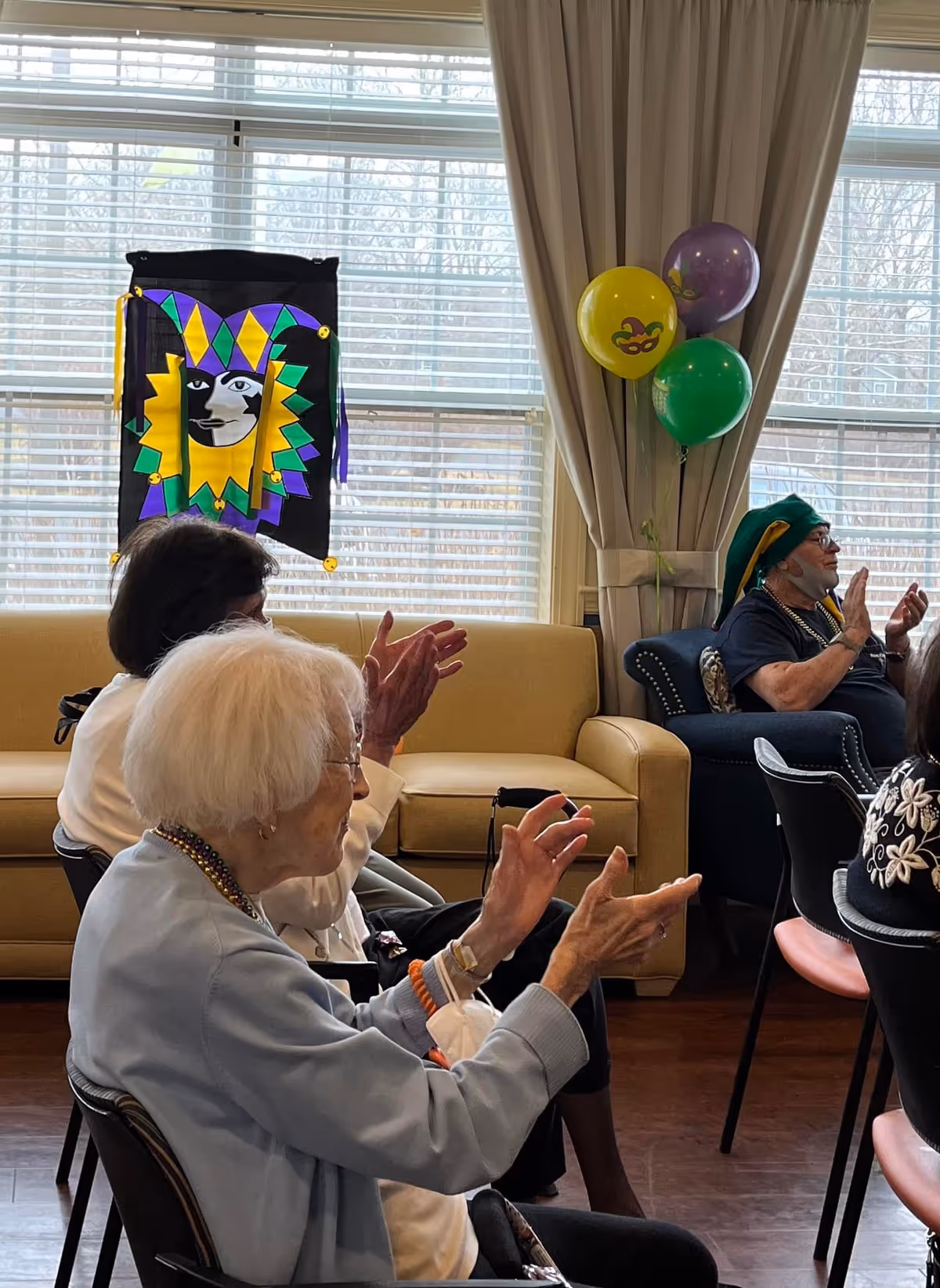 Elderly residents seated and clapping in a decorated common room with a Mardi Gras banner and balloons.