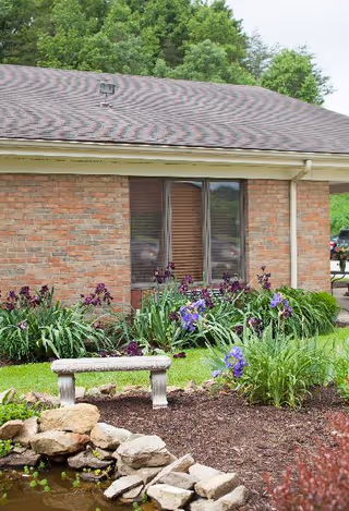 A garden area outside a brick building with a small stone bench, purple flowers, green plants, and a small pond with rocks. The building has a window with closed blinds and a sloped roof. Trees are visible in the background.