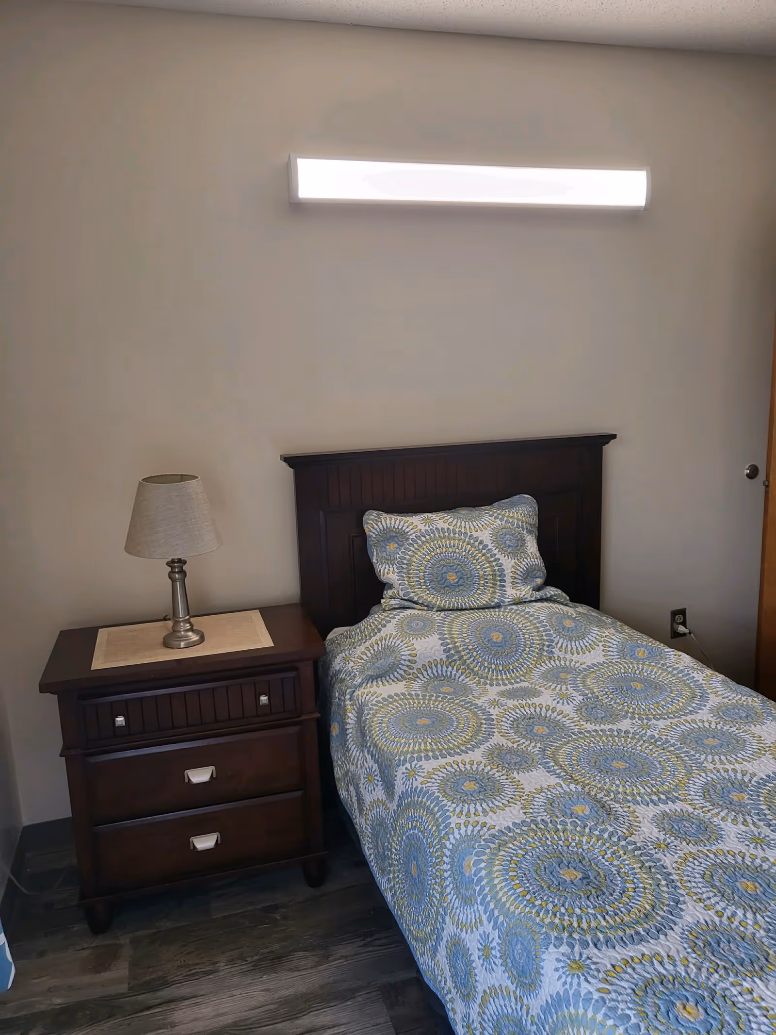 A small bedroom with a single bed featuring a patterned blue, green, and white bedspread and matching pillow. Next to the bed is a dark wooden nightstand with two drawers and a table lamp with a beige shade. A bright rectangular light fixture is mounted on the wall above the bed.